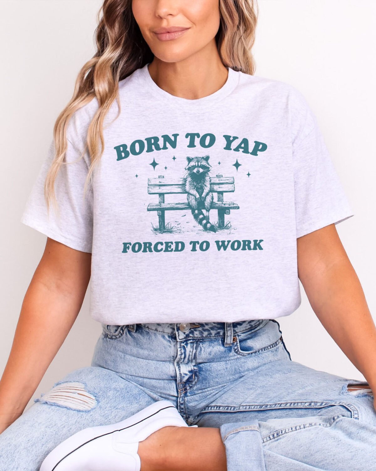 Woman wearing a Born To Yap Shirt in ash grey color with a funny vintage graphic of a raccoon sitting on a bench