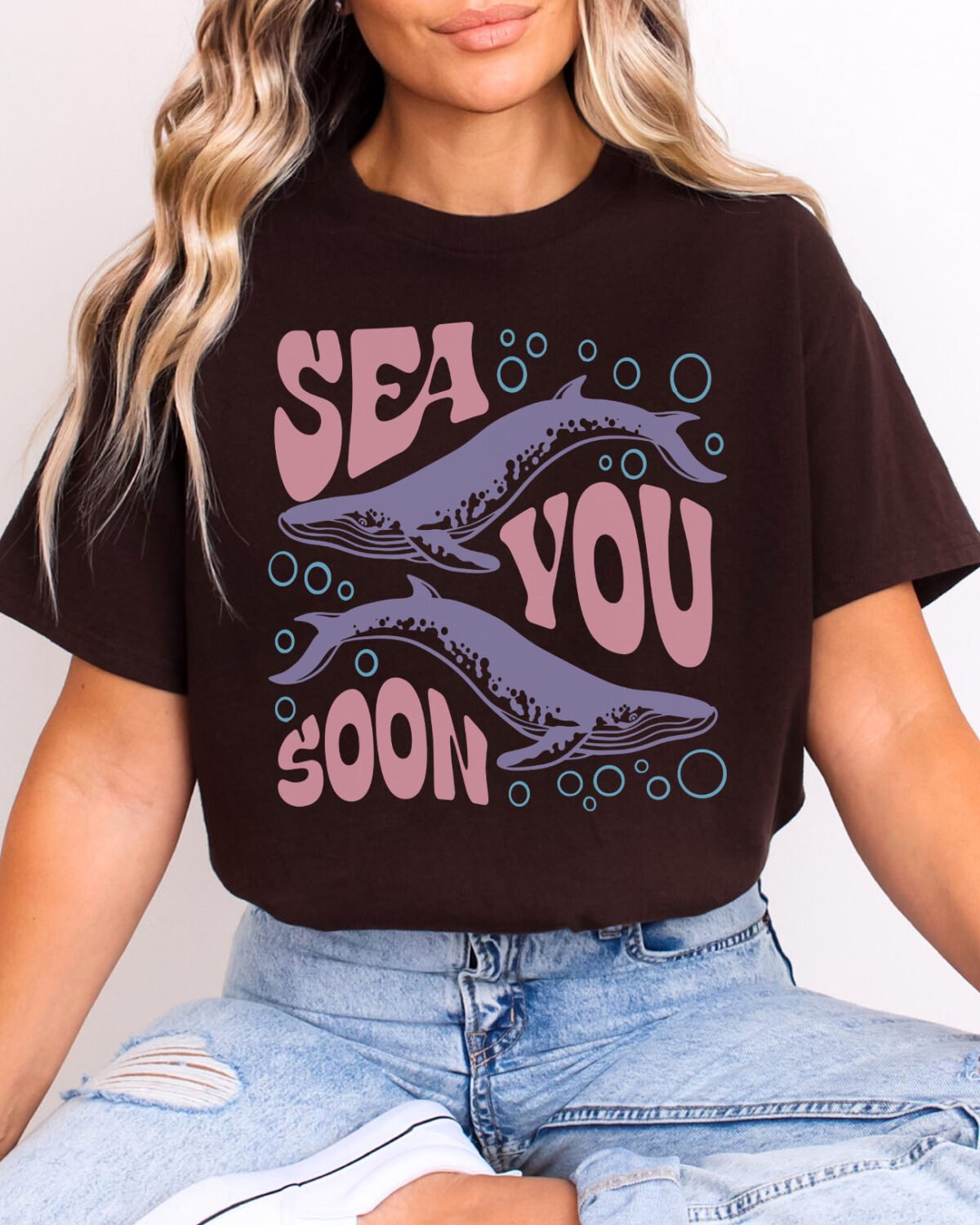 Woman wearing a Sea You Soon Shirt in dark chocolate color. The design on the shirt features a retro ocean whale graphic.